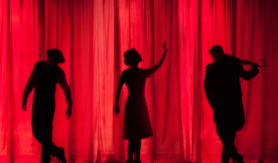 Three actors in silhouette in front of a red theatre curtain
